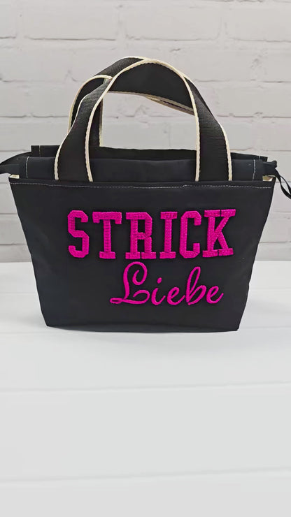 Projekttasche Alva S aus schwarzem Canvas mit erhabenem 3D-Stickzug Strickliebe in Pink, Henkeln und Kordelzugkragen. Handgenäht in Bayern von Nadelmikado.