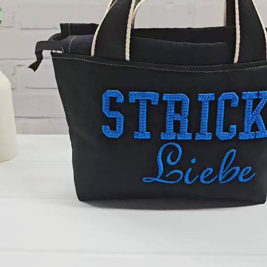 Projekttasche Alva S aus schwarzem Canvas mit blauer 3D-Stickerei Strickliebe, zwei Henkeln und schwarzem Kordelzugkragen. Handgenäht in Bayern von Nadelmikado.