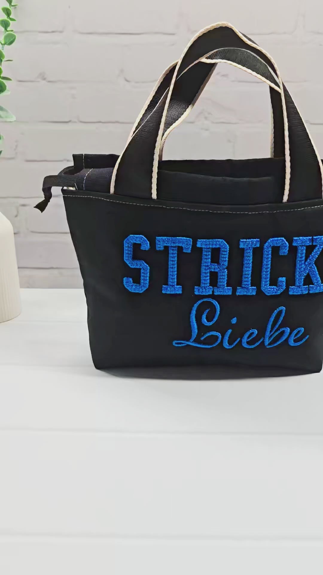 Projekttasche Alva S aus schwarzem Canvas mit blauer 3D-Stickerei Strickliebe, zwei Henkeln und schwarzem Kordelzugkragen. Handgenäht in Bayern von Nadelmikado.