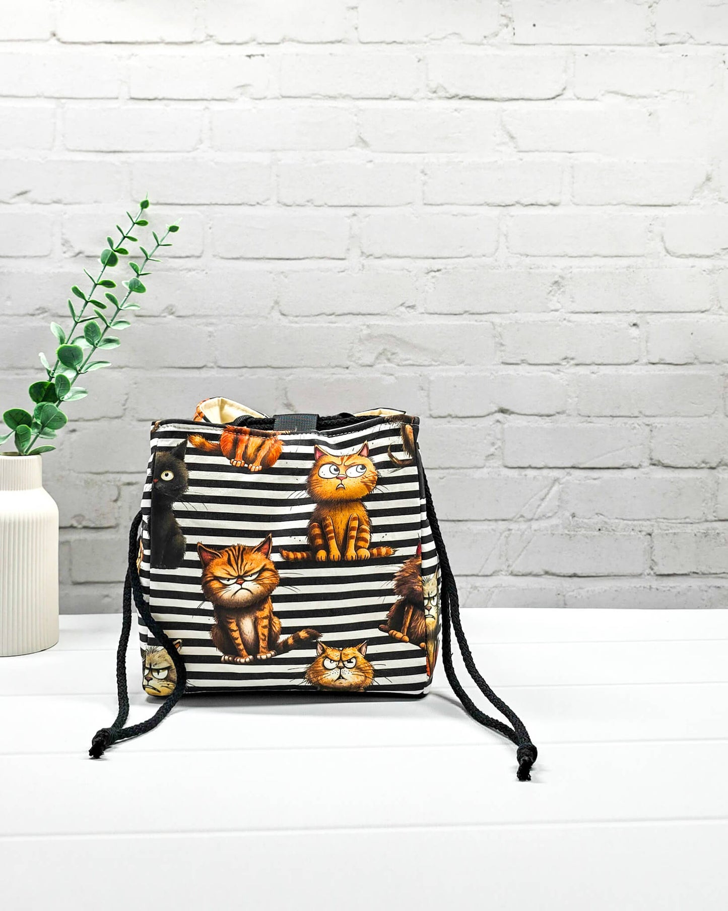 Mittlerer Projektbeutel Aska M als Rice Bag mit schwarz-weißem Streifenmuster, Grumpy-Cat-Motiven und seitlichen schwarzen Kordeln zum Zuziehen. Handgenäht in Bayern von Nadelmikado.