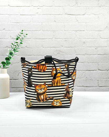 Mittlerer Projektbeutel Aska M als Rice Bag mit schwarz-weißem Streifenmuster, Grumpy-Cat-Motiven und seitlichen schwarzen Kordeln zum Zuziehen. Handgenäht in Bayern von Nadelmikado.