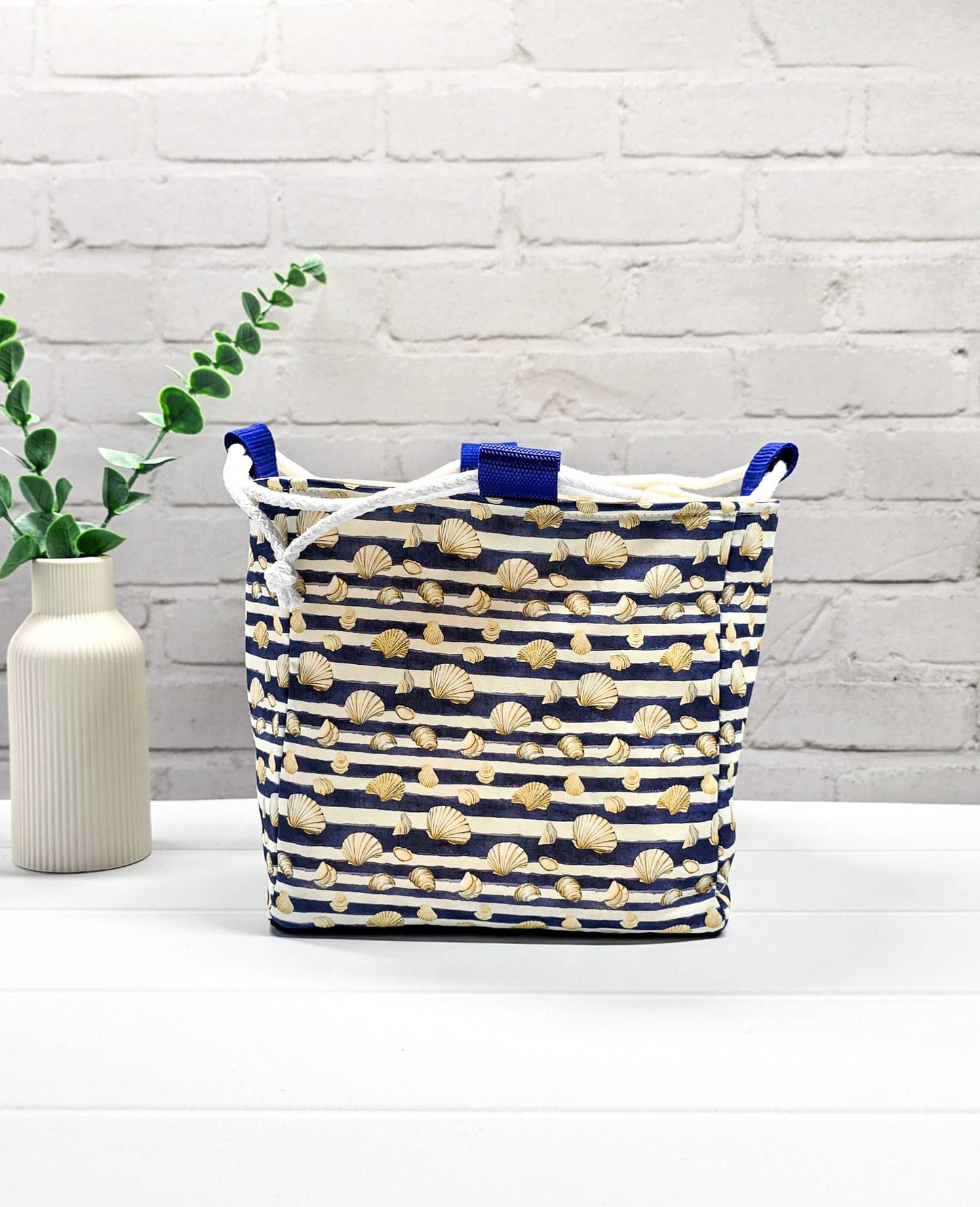 Projektbeutel Aska M aus blau-weiß quergestreiftem Stoff mit kleinen Muschelmotiven, zwei seitlich herausgeführten, hellen Kordeln und einer blauen Schlaufe an der Oberkante. Rice Bag. Handgenäht in Bayern von Nadelmikado. 