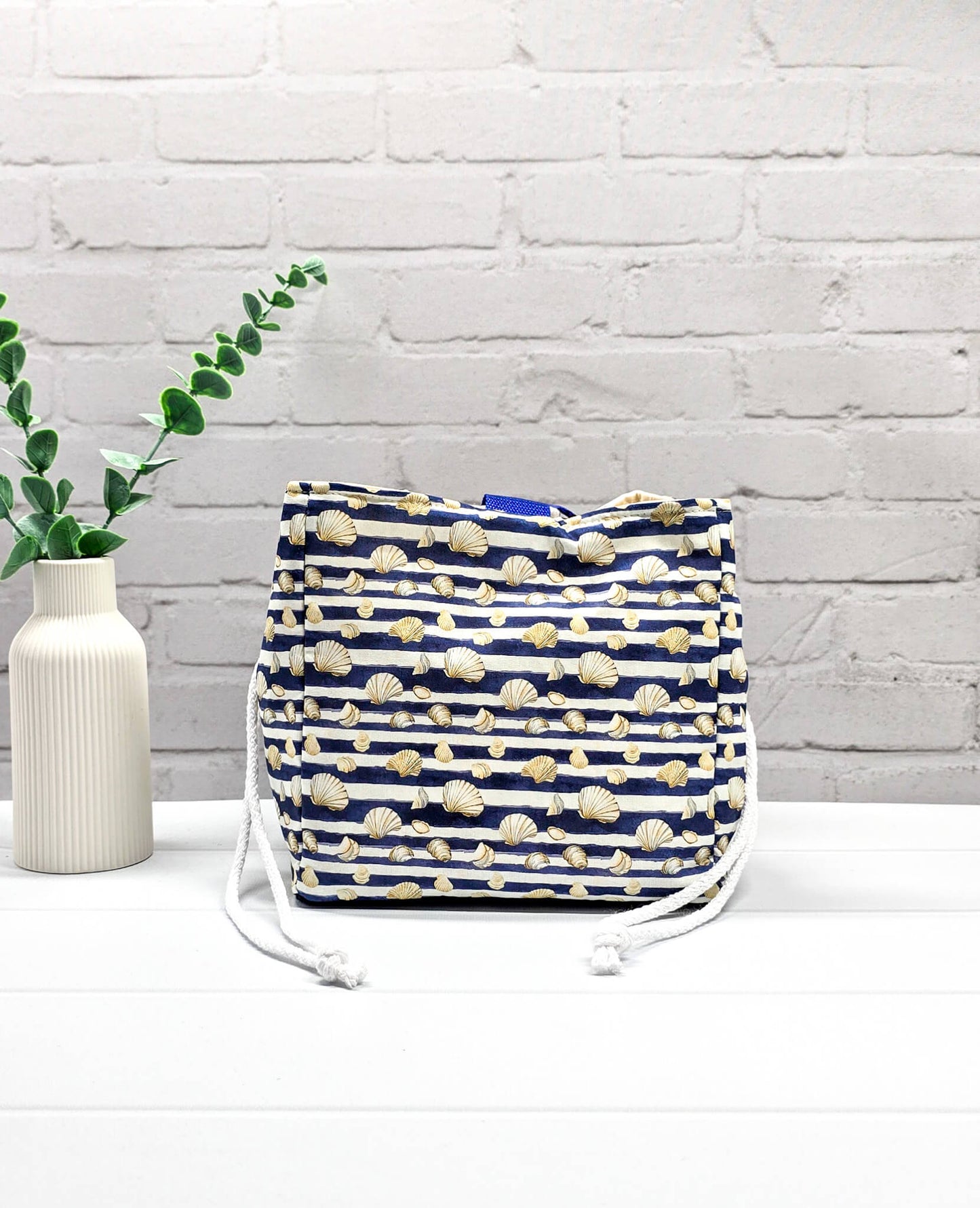 Projektbeutel Aska M aus blau-weiß quergestreiftem Stoff mit kleinen Muschelmotiven, zwei seitlich herausgeführten, hellen Kordeln und einer blauen Schlaufe an der Oberkante. Rice Bag. Handgenäht in Bayern von Nadelmikado. 