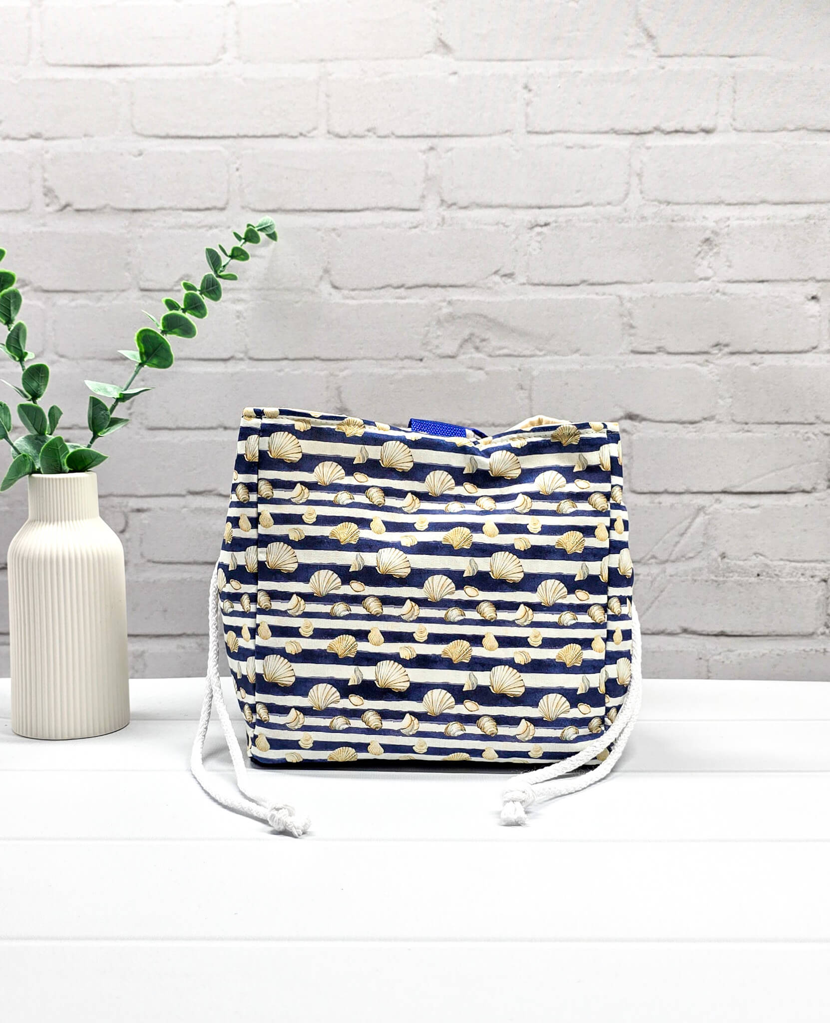 Projektbeutel Aska M aus blau-weiß quergestreiftem Stoff mit kleinen Muschelmotiven, zwei seitlich herausgeführten, hellen Kordeln und einer blauen Schlaufe an der Oberkante. Rice Bag. Handgenäht in Bayern von Nadelmikado. 