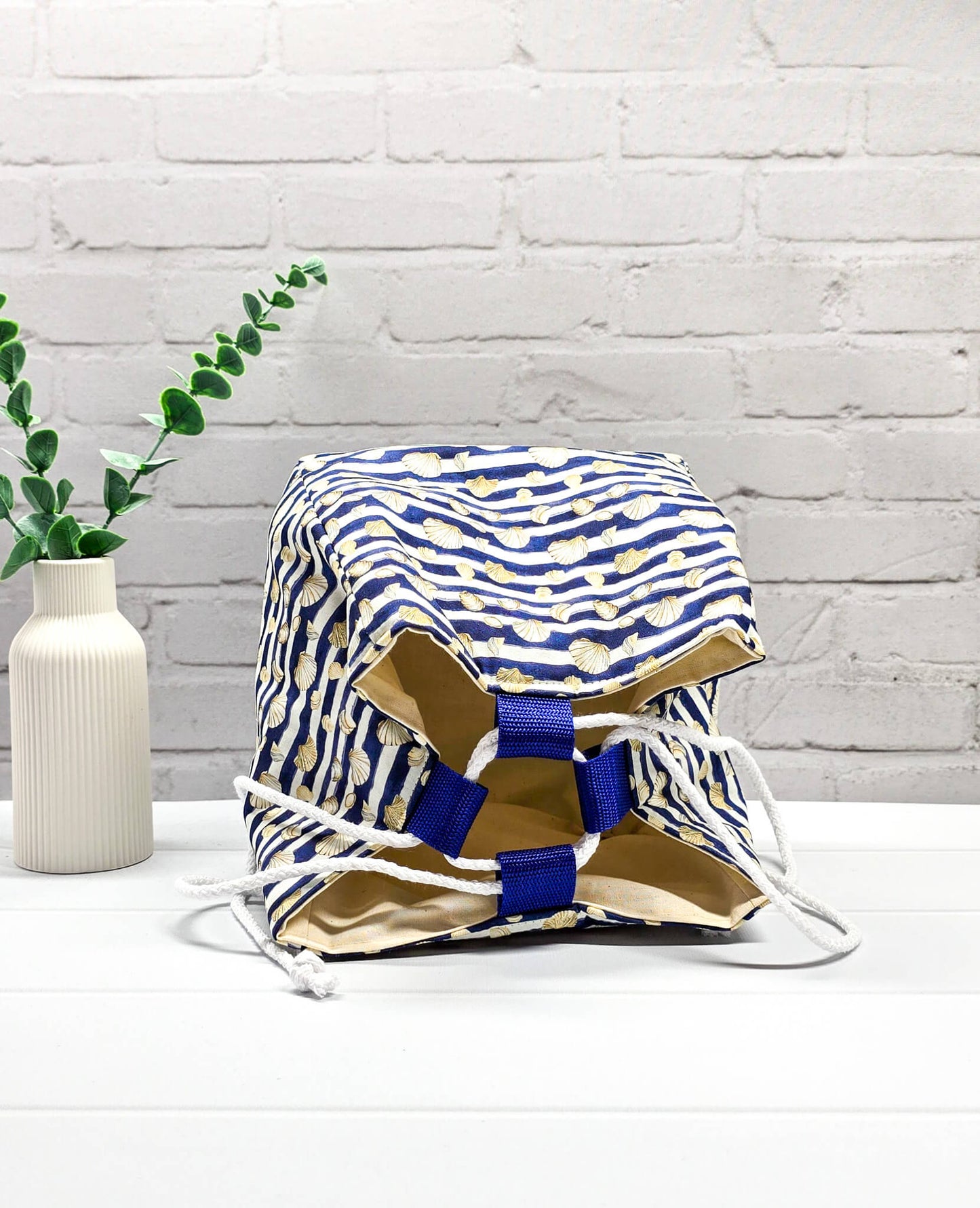 Projektbeutel Aska M aus blau-weiß quergestreiftem Stoff mit kleinen Muschelmotiven, zwei seitlich herausgeführten, hellen Kordeln und einer blauen Schlaufe an der Oberkante. Rice Bag. Handgenäht in Bayern von Nadelmikado. 