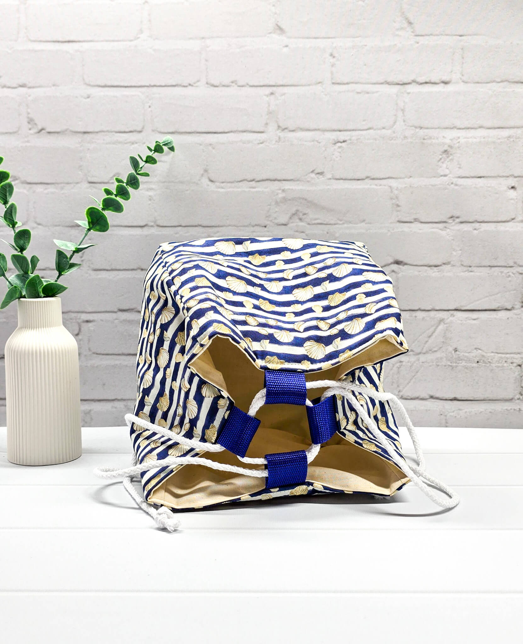 Projektbeutel Aska M aus blau-weiß quergestreiftem Stoff mit kleinen Muschelmotiven, zwei seitlich herausgeführten, hellen Kordeln und einer blauen Schlaufe an der Oberkante. Rice Bag. Handgenäht in Bayern von Nadelmikado. 