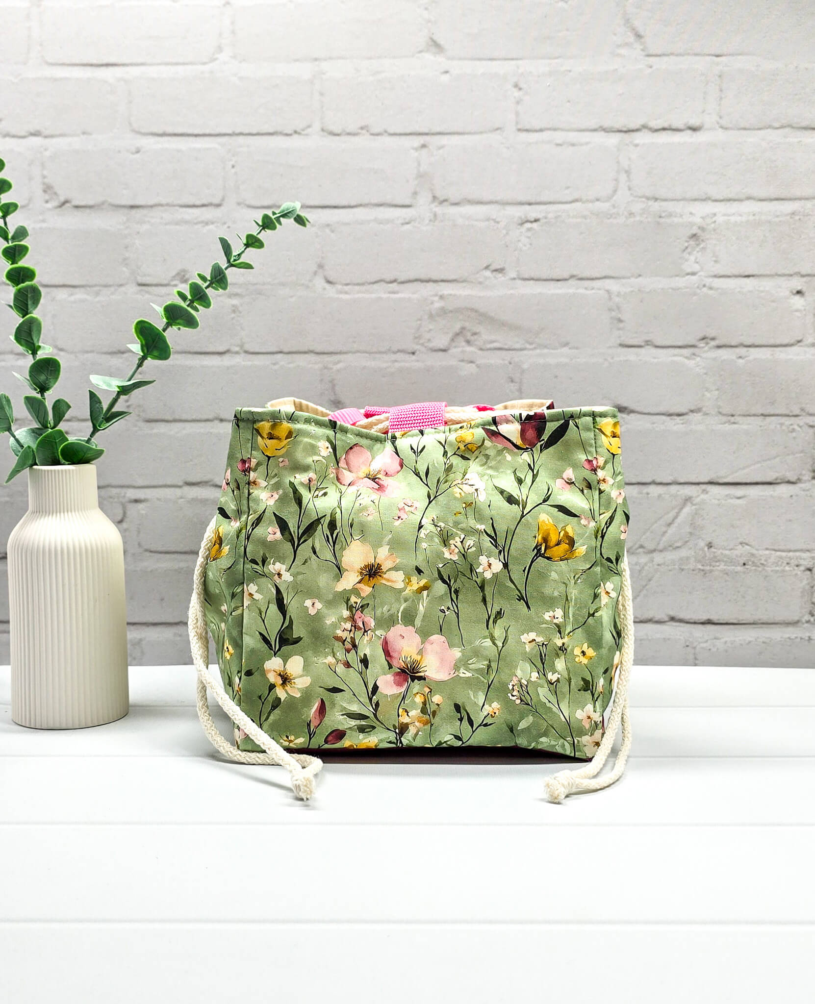 Projektbeutel Aska M aus salbeigrünem Stoff mit buntem Wildblumenmuster, zwei cremeweißen Kordeln zum Zuziehen und einer rosa Schlaufe an der Oberkante. Handgenäht in Bayern von Nadelmikado. 