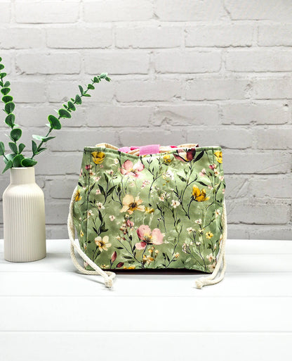 Projektbeutel Aska M aus salbeigrünem Stoff mit buntem Wildblumenmuster, zwei cremeweißen Kordeln zum Zuziehen und einer rosa Schlaufe an der Oberkante. Handgenäht in Bayern von Nadelmikado. 