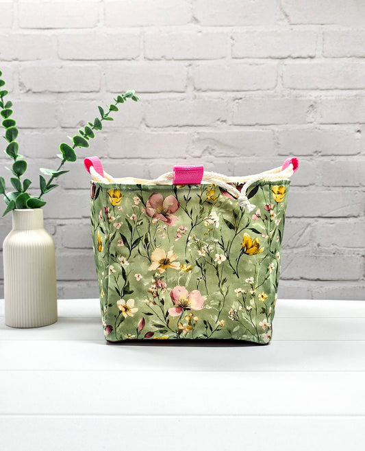 Projektbeutel Aska M aus salbeigrünem Stoff mit buntem Wildblumenmuster, zwei cremeweißen Kordeln zum Zuziehen und einer rosa Schlaufe an der Oberkante. Handgenäht in Bayern von Nadelmikado. 