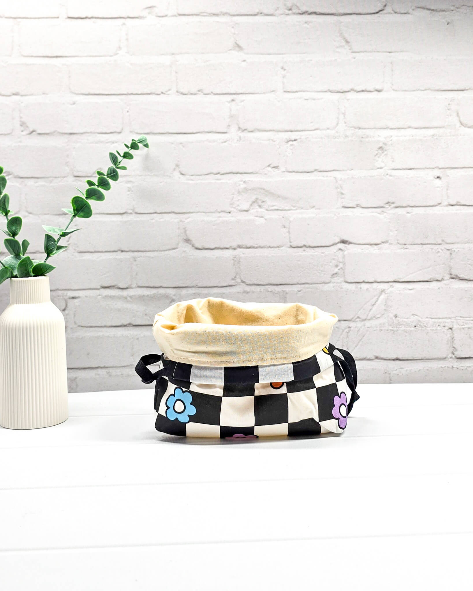 Projektbeutel Lyra bedruckt mit einem schwarz-weißen Schachbrettmuster und bunten, einfachen Blumenblüten sowie schwarzen Kordelzügen. Handgenäht in Bayern von Nadelmikado. 