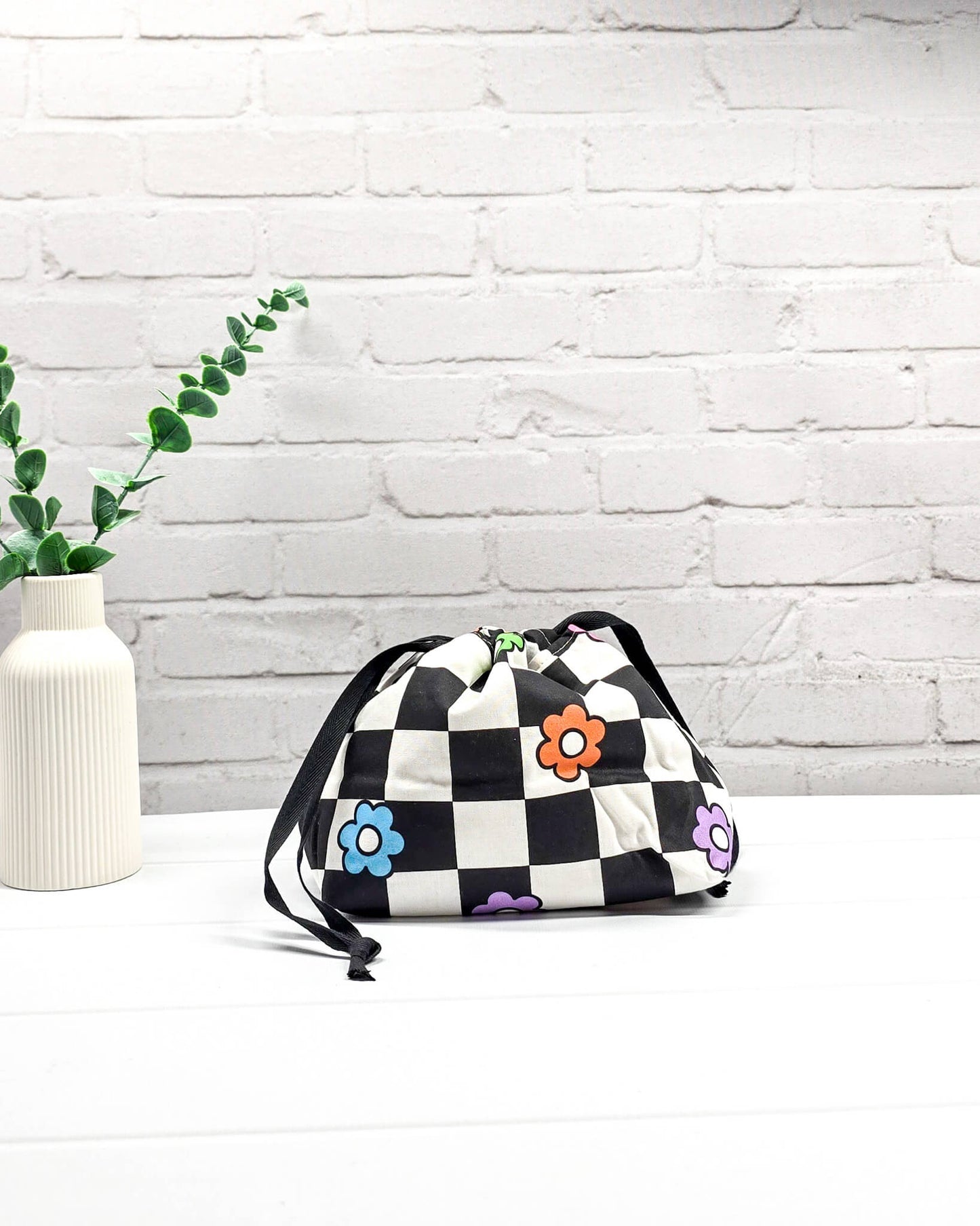 Projektbeutel Lyra bedruckt mit einem schwarz-weißen Schachbrettmuster und bunten, einfachen Blumenblüten sowie schwarzen Kordelzügen. Handgenäht in Bayern von Nadelmikado. 