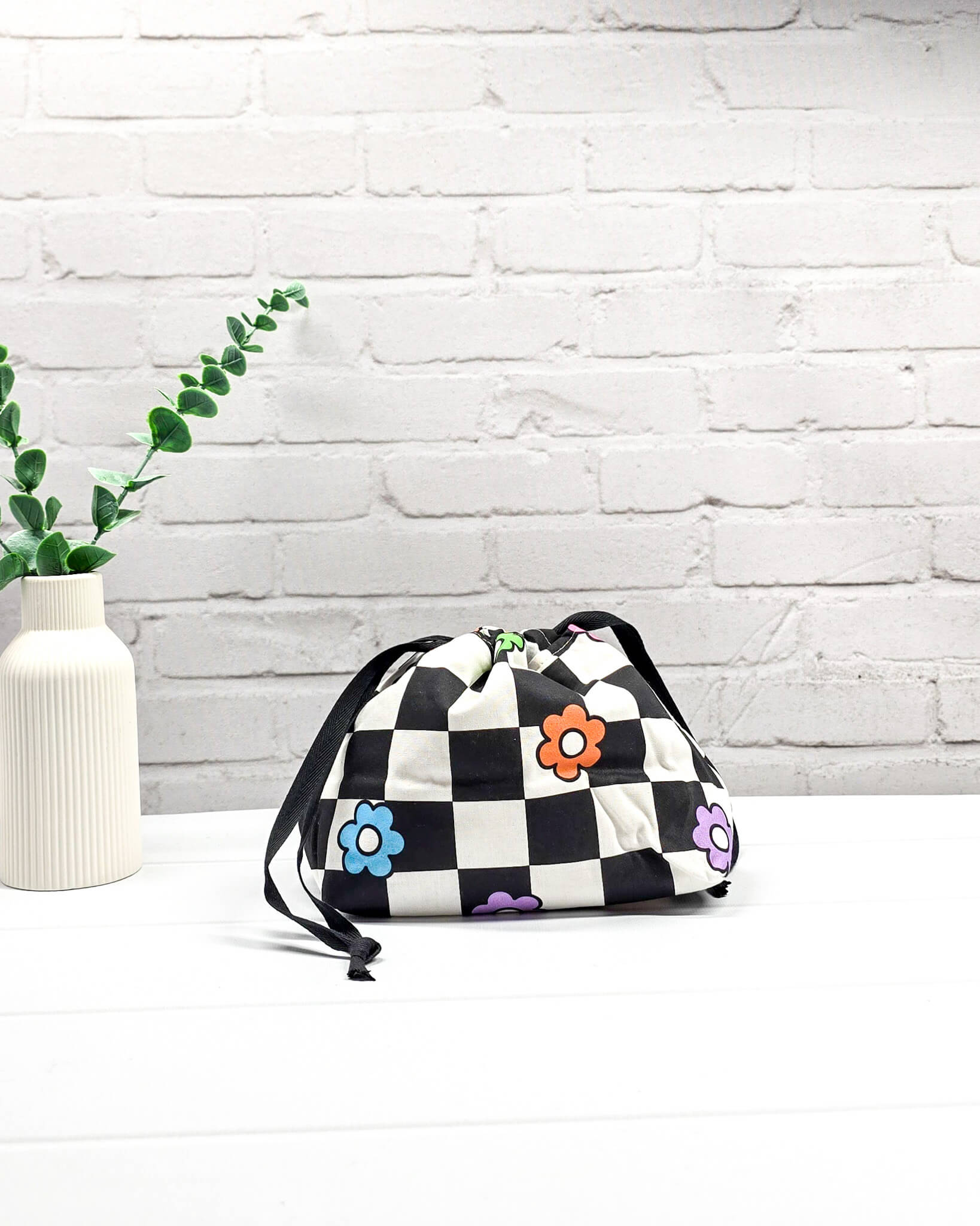 Projektbeutel Lyra bedruckt mit einem schwarz-weißen Schachbrettmuster und bunten, einfachen Blumenblüten sowie schwarzen Kordelzügen. Handgenäht in Bayern von Nadelmikado. 