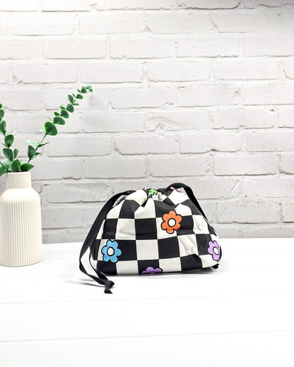 Projektbeutel Lyra bedruckt mit einem schwarz-weißen Schachbrettmuster und bunten, einfachen Blumenblüten sowie schwarzen Kordelzügen. Handgenäht in Bayern von Nadelmikado. 