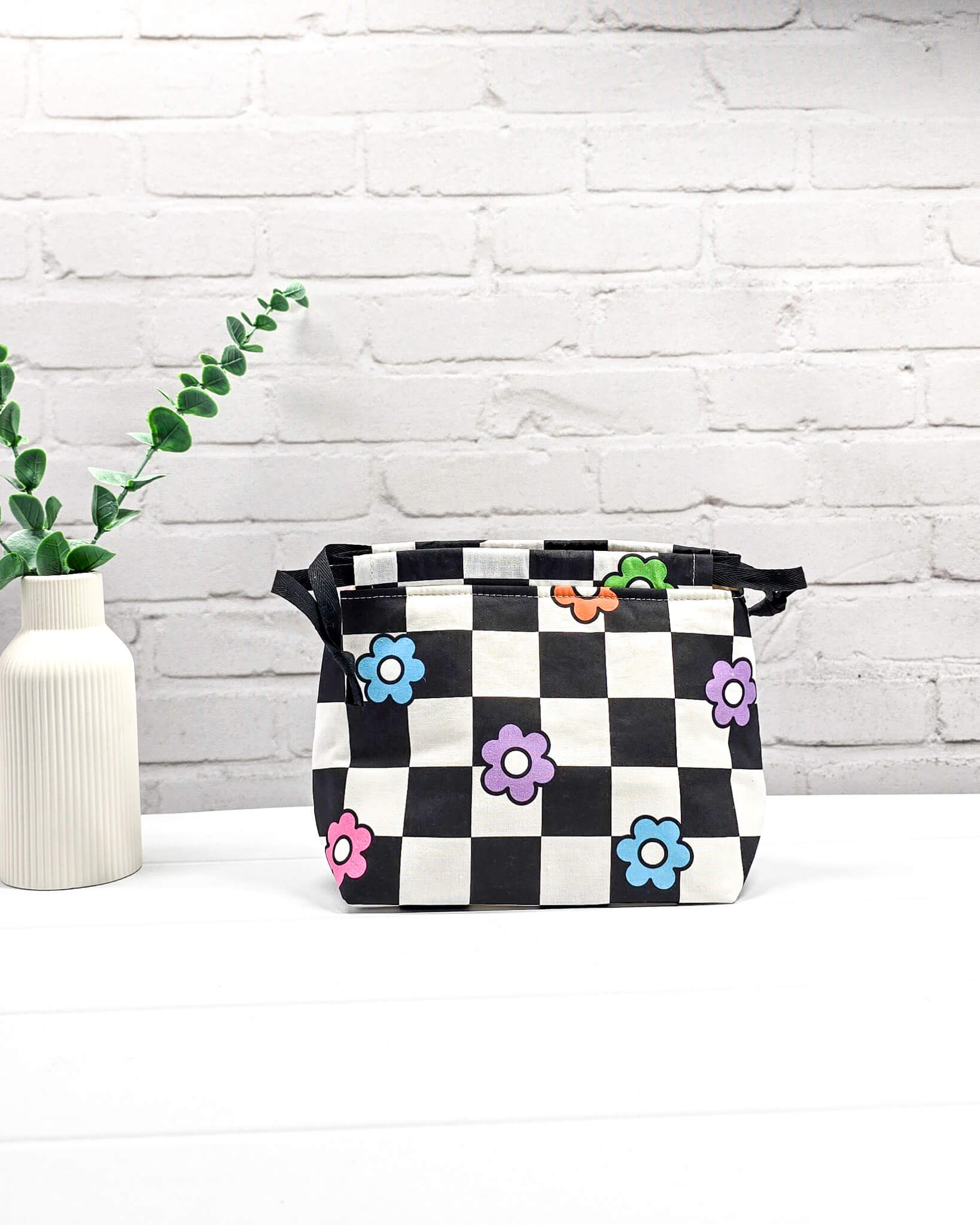 Projektbeutel Lyra bedruckt mit einem schwarz-weißen Schachbrettmuster und bunten, einfachen Blumenblüten sowie schwarzen Kordelzügen. Handgenäht in Bayern von Nadelmikado. 
