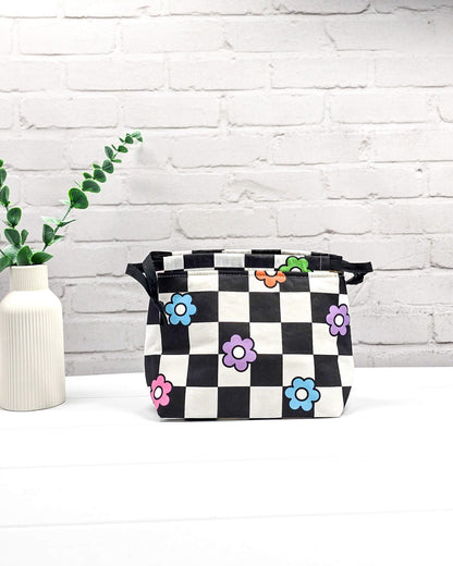 Projektbeutel Lyra bedruckt mit einem schwarz-weißen Schachbrettmuster und bunten, einfachen Blumenblüten sowie schwarzen Kordelzügen. Handgenäht in Bayern von Nadelmikado. 