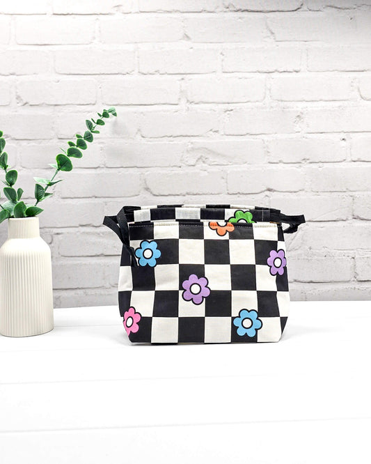 Projektbeutel Lyra bedruckt mit einem schwarz-weißen Schachbrettmuster und bunten, einfachen Blumenblüten sowie schwarzen Kordelzügen. Handgenäht in Bayern von Nadelmikado. 