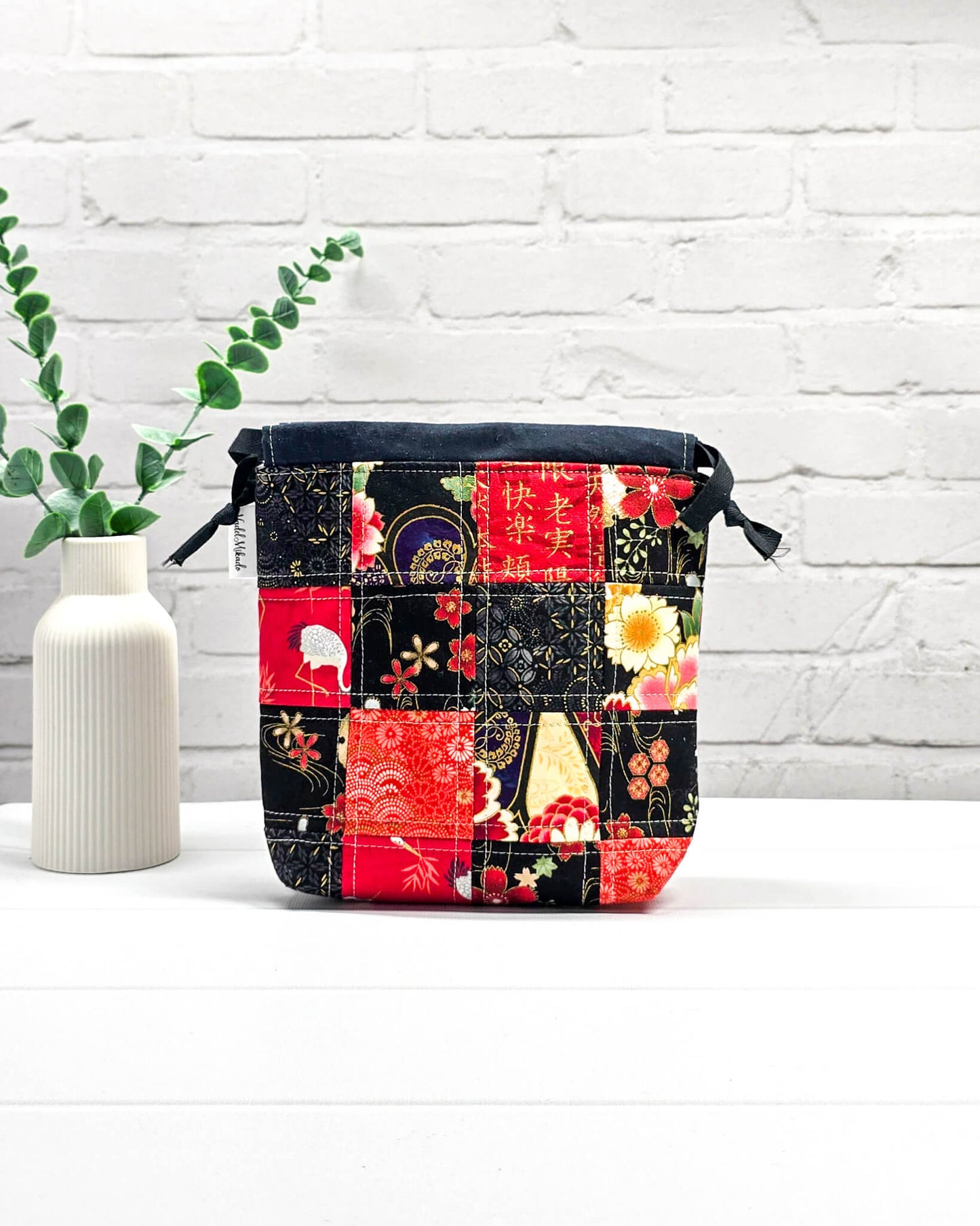 Projektbeutel Tindra bedruckt mit einem roten und schwarzen Patchwork-Muster aus Kranichen und Blumenblüten sowie schwarzen Kordelzügen. Handgenäht in Bayern von Nadelmikado. 