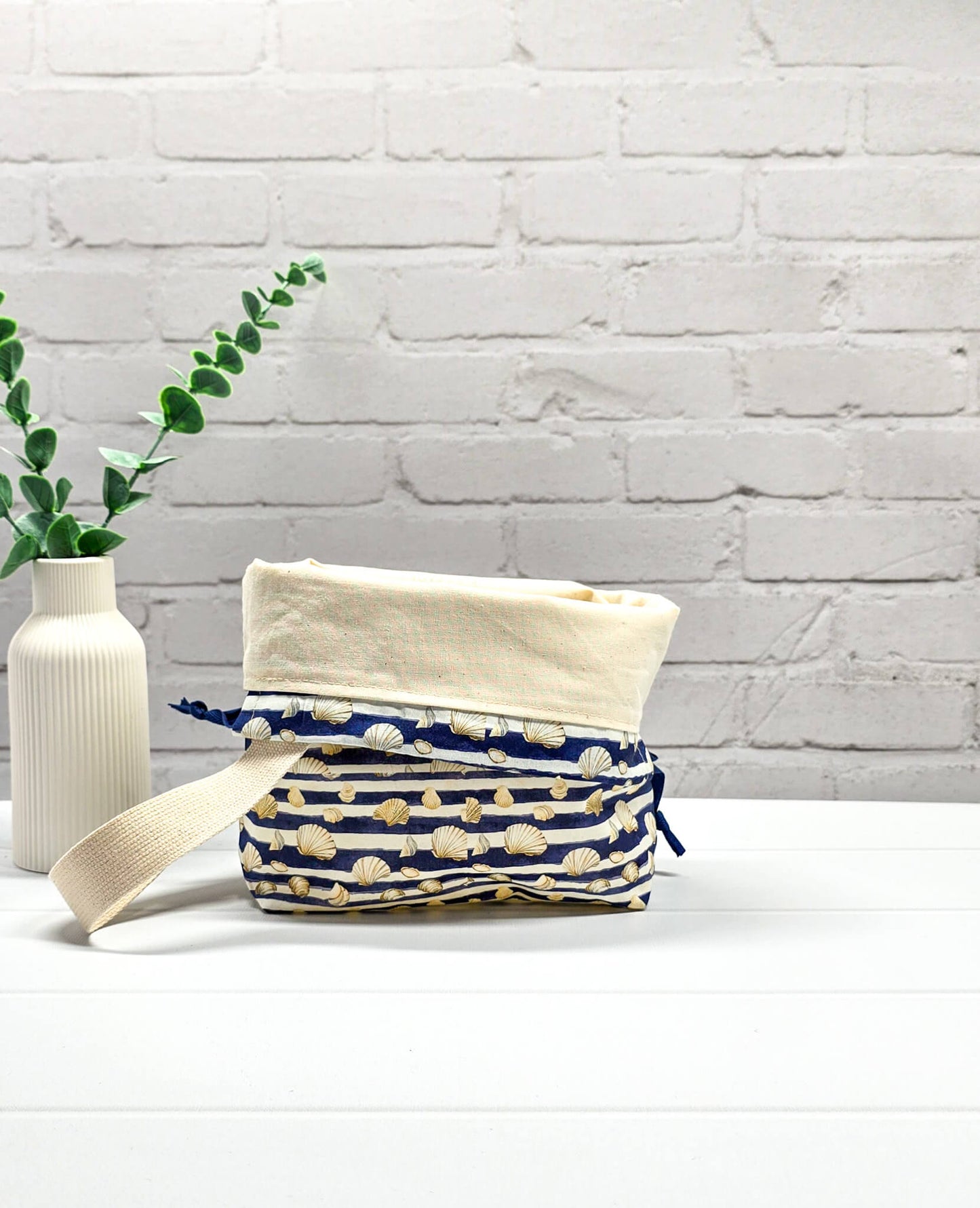 Projektbeutel Tuva S aus blau-weiß quergestreiftem Stoff mit kleinen Muschelmotiven, einer hellen Gurtband-Schlaufe und dunkelblauem Kordelzug. Handgenäht in Bayern von Nadelmikado. 