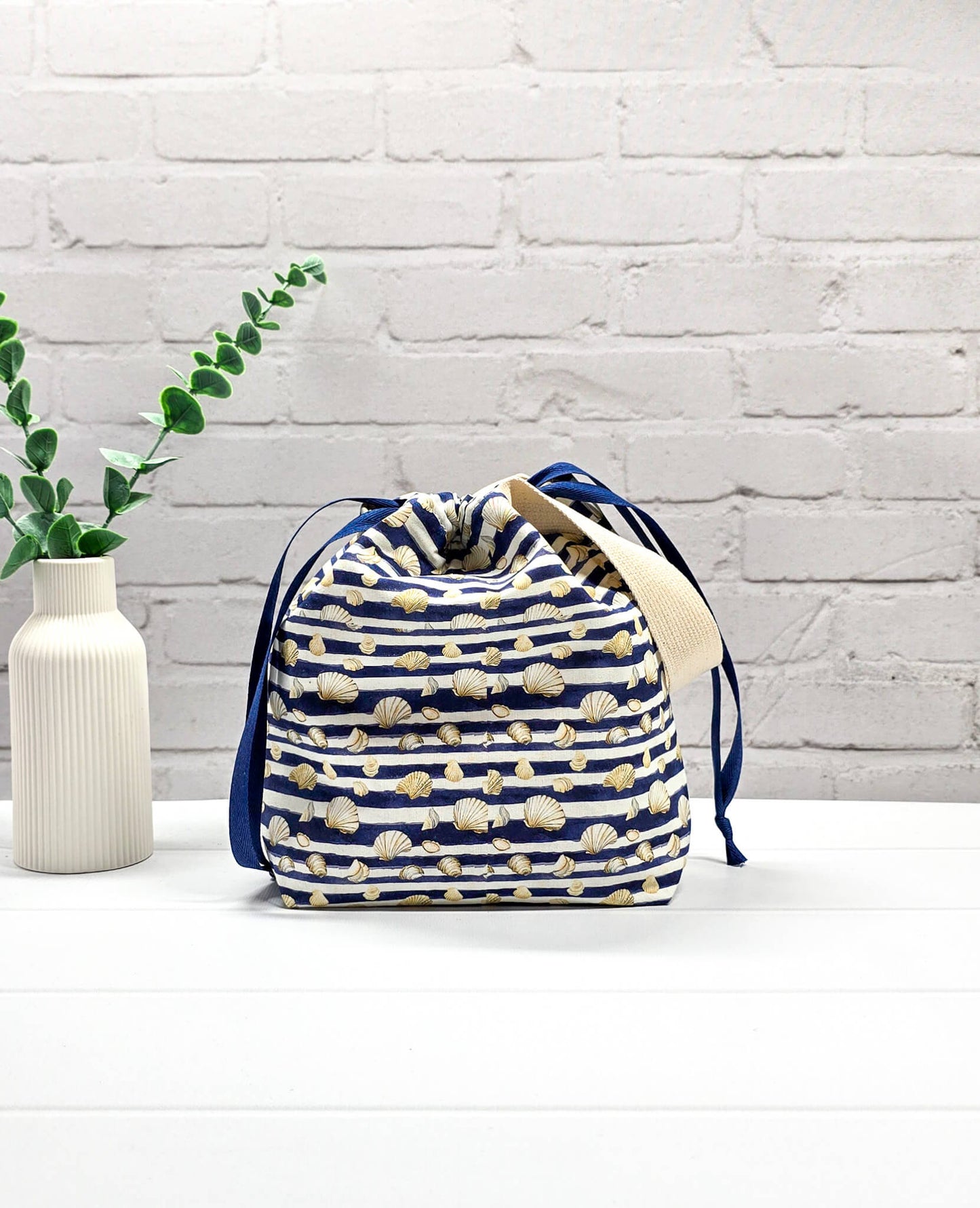 Projektbeutel Tuva S aus blau-weiß quergestreiftem Stoff mit kleinen Muschelmotiven, einer hellen Gurtband-Schlaufe und dunkelblauem Kordelzug. Handgenäht in Bayern von Nadelmikado. 