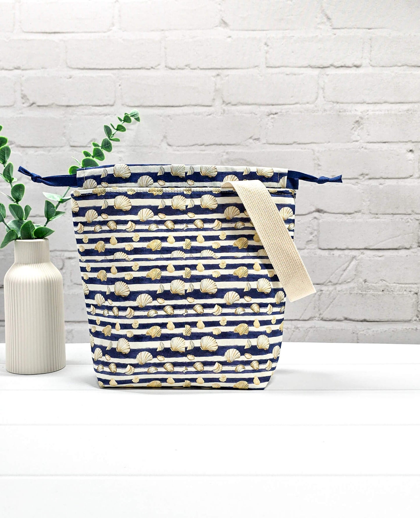 Projektbeutel Tuva S aus blau-weiß quergestreiftem Stoff mit kleinen Muschelmotiven, einer hellen Gurtband-Schlaufe und dunkelblauem Kordelzug. Handgenäht in Bayern von Nadelmikado. 