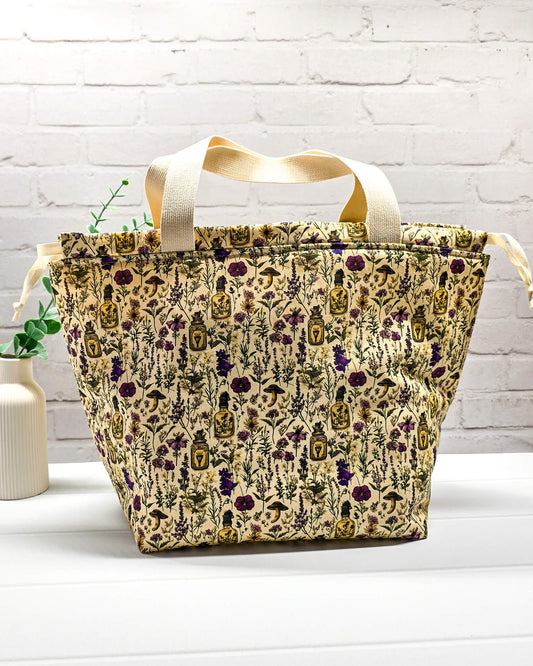 Große Projekttasche Alva L mit zwei hellen Tragehenkeln, botanischem Kräuterküche-Muster auf beigem Grund und hellen Zugkordeln. Handgenäht in Bayern von Nadelmikado.