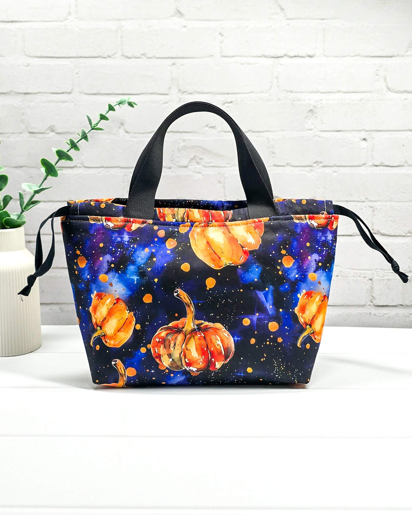 Projekttasche Alva S mit orangefarbenen Kürbissen auf dunkelblauem Galaxie-Sternenhimmel-Stoff und schwarzen Henkeln. Handgenäht in Bayern von Nadelmikado