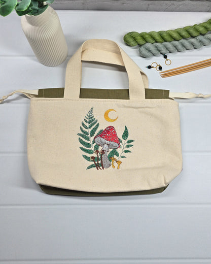 Projekttasche Alva S aus hellem Canvas mit Stickmotiv Fliegenpilz Farn und Mondsichel, Henkeln und olivgrünem Kordelzug. Handgenäht in Bayern von Nadelmikado.