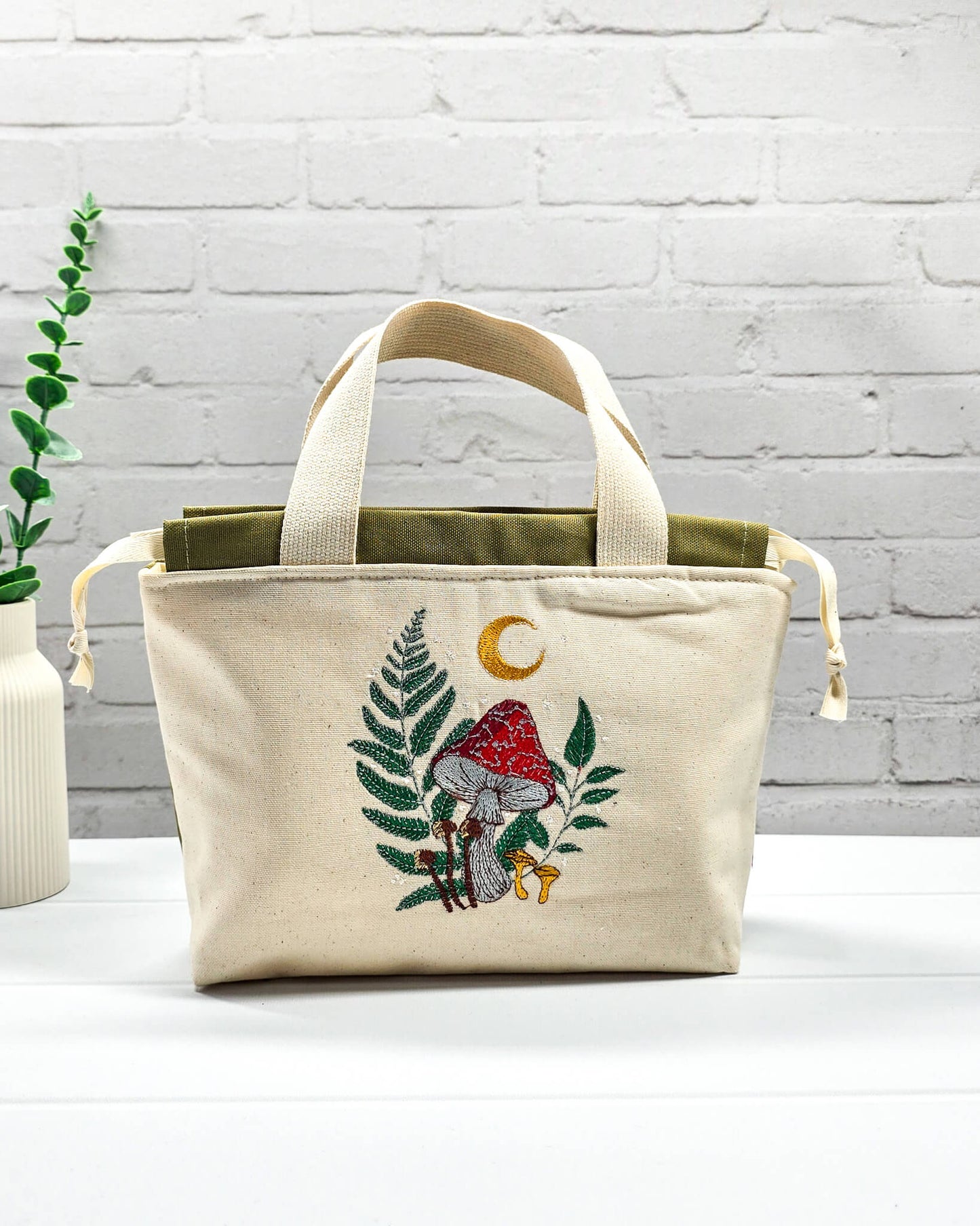 Projekttasche Alva S aus hellem Canvas mit Stickmotiv Fliegenpilz Farn und Mondsichel, Henkeln und olivgrünem Kordelzug. Handgenäht in Bayern von Nadelmikado.