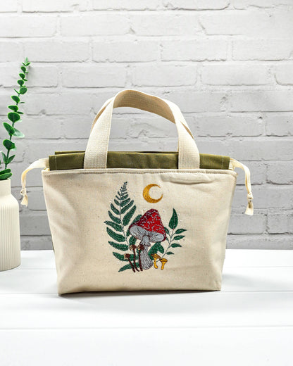 Projekttasche Alva S aus hellem Canvas mit Stickmotiv Fliegenpilz Farn und Mondsichel, Henkeln und olivgrünem Kordelzug. Handgenäht in Bayern von Nadelmikado.