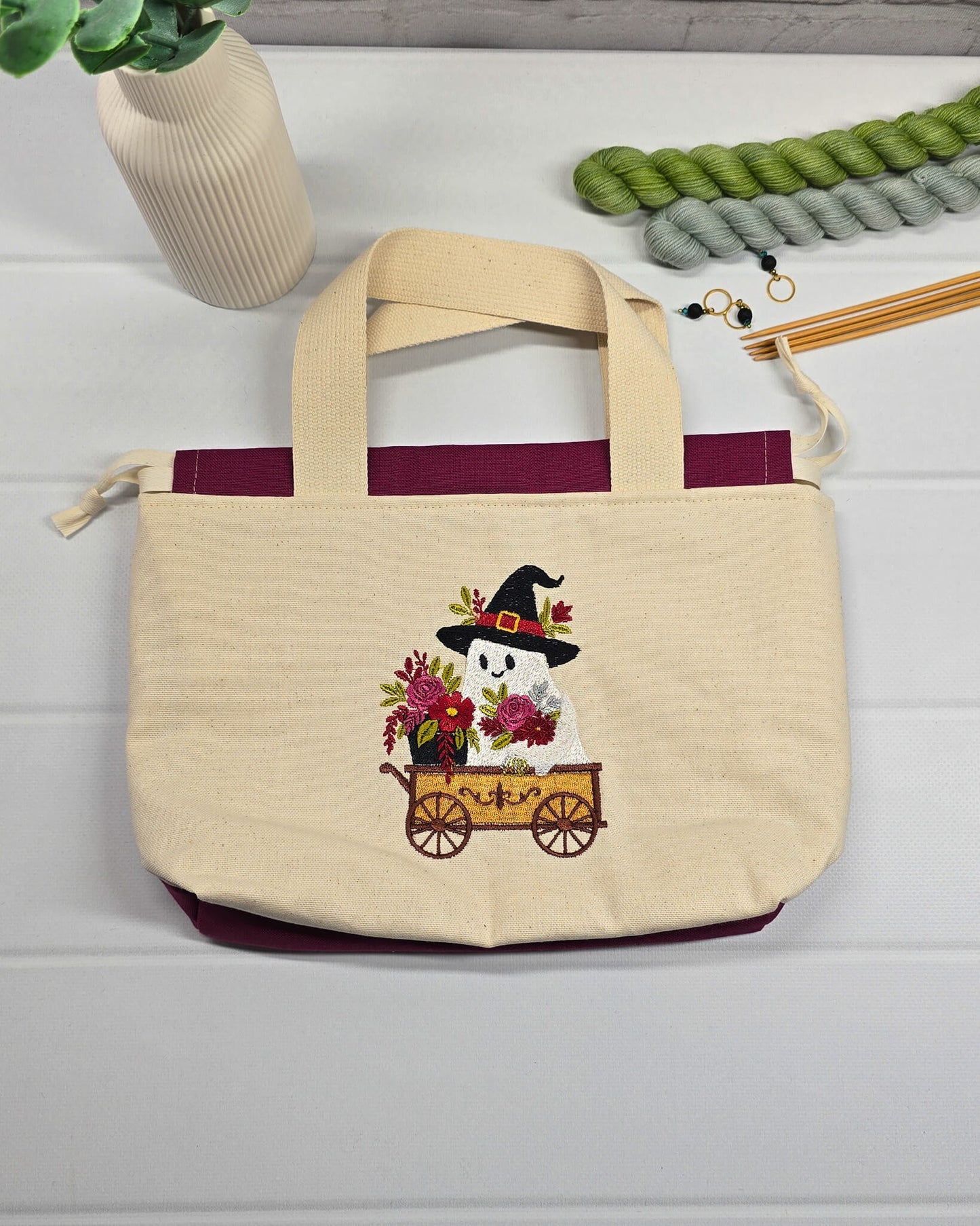Projekttasche Alva S aus hellem Canvas mit gesticktem Geist im Bollerwagen, Henkeln und bordeauxrotem Kordelzug. Handgenäht in Bayern von Nadelmikado.