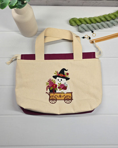 Projekttasche Alva S aus hellem Canvas mit gesticktem Geist im Bollerwagen, Henkeln und bordeauxrotem Kordelzug. Handgenäht in Bayern von Nadelmikado.