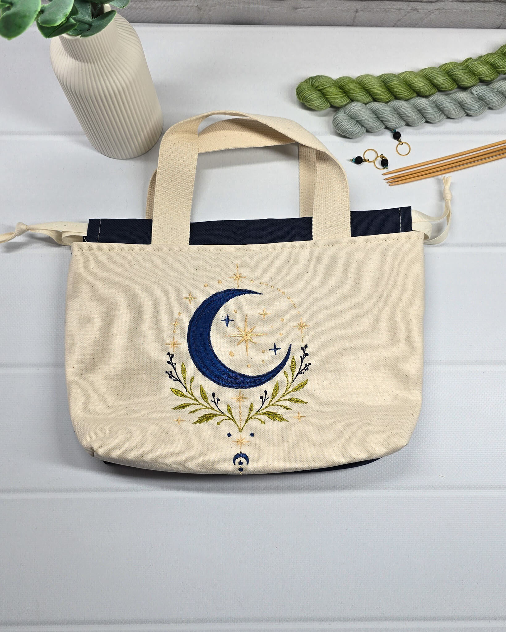Projekttasche Alva S aus hellem Canvas mit Stickmotiv blaue Mondsichel und Sterne, Henkeln und dunkelblauem Kordelzug. Handgenäht in Bayern von Nadelmikado.