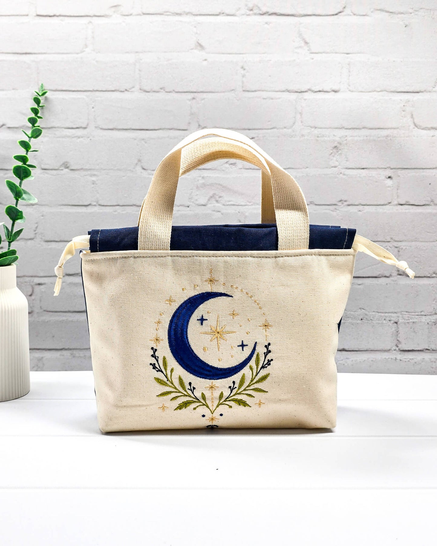 Projekttasche Alva S aus hellem Canvas mit Stickmotiv blaue Mondsichel und Sterne, Henkeln und dunkelblauem Kordelzug. Handgenäht in Bayern von Nadelmikado.
