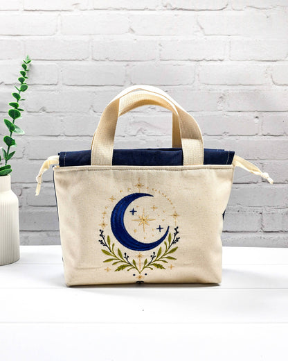Projekttasche Alva S aus hellem Canvas mit Stickmotiv blaue Mondsichel und Sterne, Henkeln und dunkelblauem Kordelzug. Handgenäht in Bayern von Nadelmikado.
