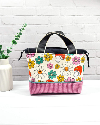 Handgemachte Projekttasche Alva in Größe S mit zwei Tragegriffen, bunten Retro-Pilzen und Blumen auf hellem Stoff und einem Taschenboden aus altrosa Breitcord. Handgenäht in Bayern von Nadelmikado. 