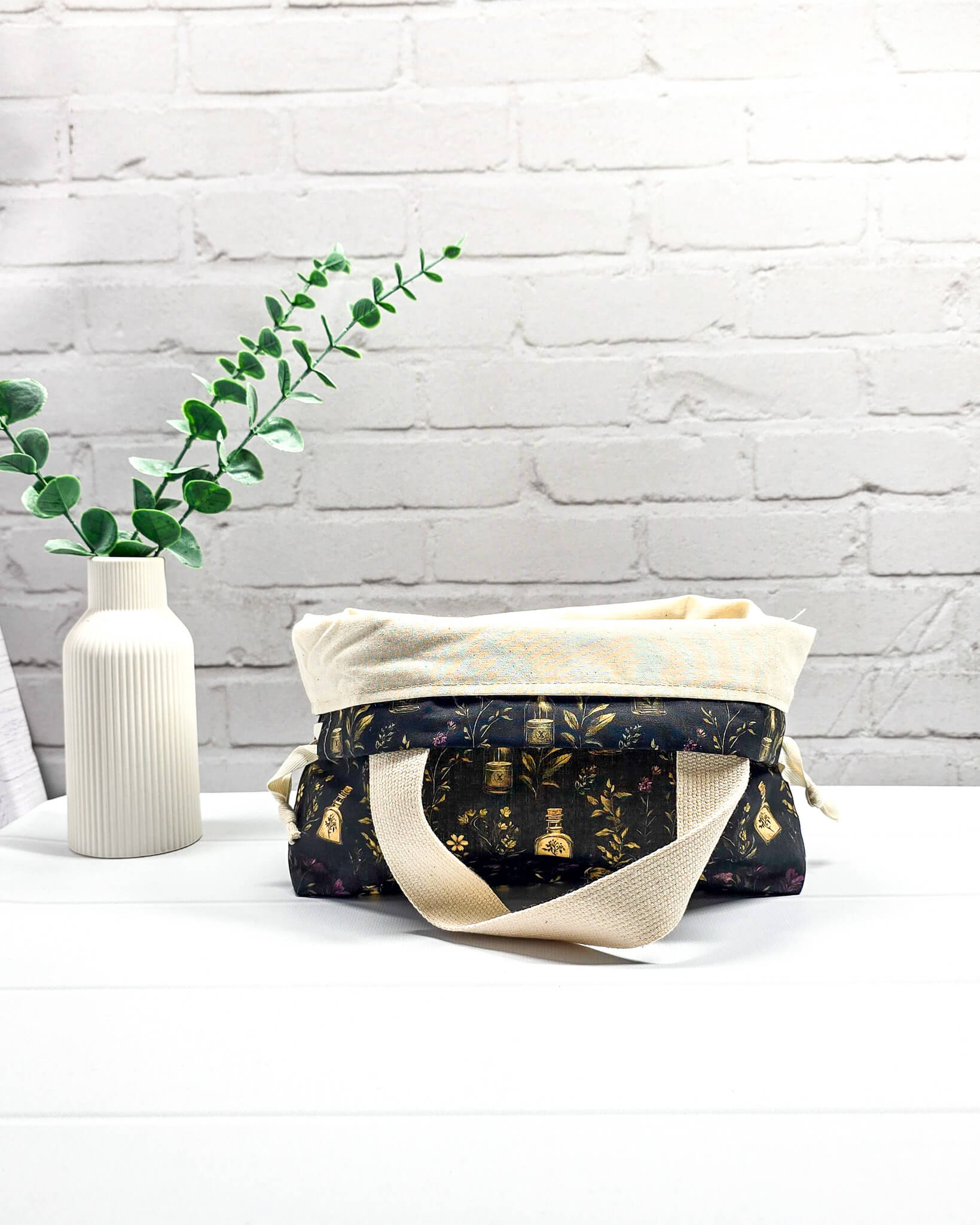 Projekttasche Alva S Secret Garden mit zwei hellen Griffen und dunklem botanischem Muster mit Blüten und kleinen goldenen Fläschchen. Handgenäht in Bayern von NadelMikado.