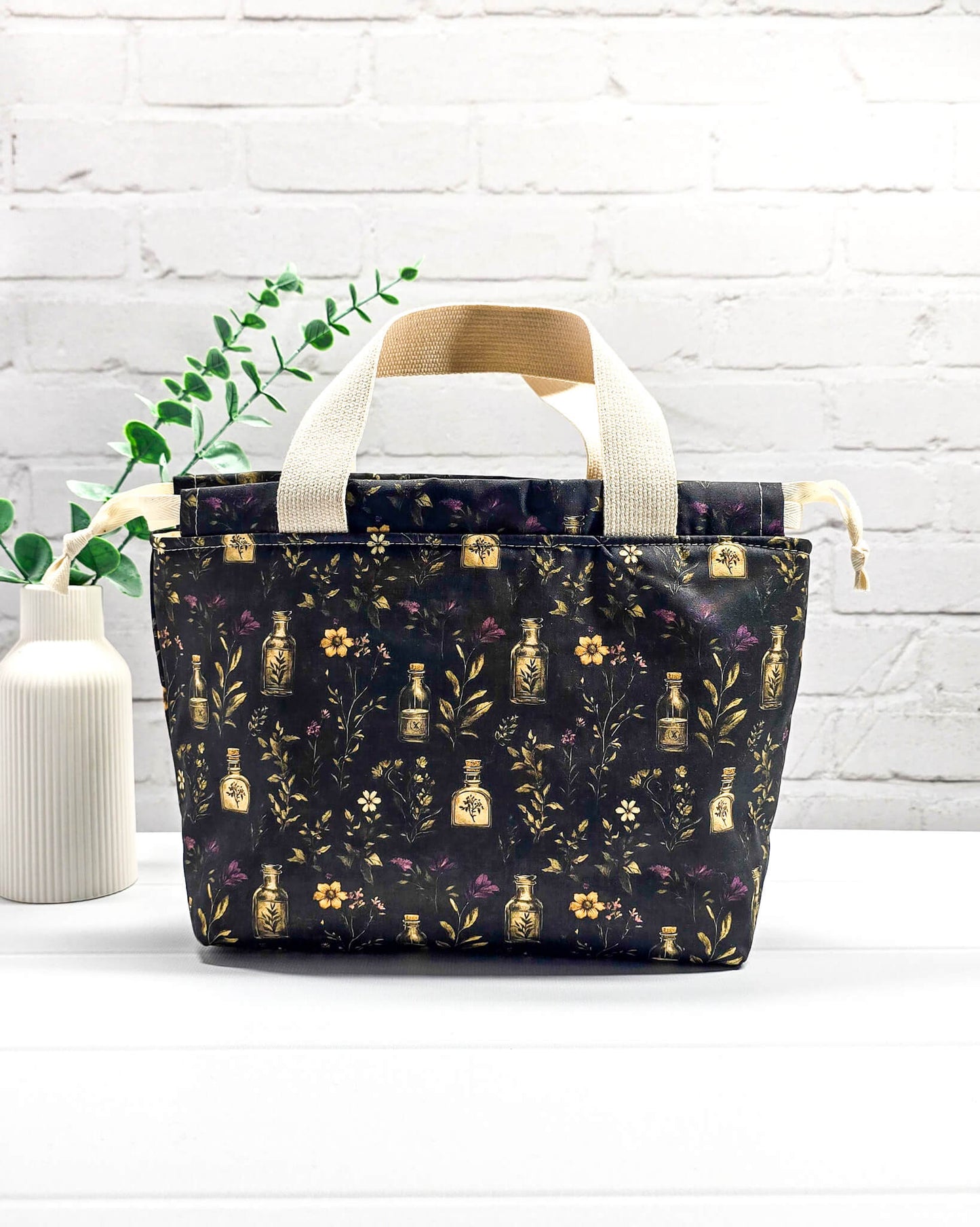 Projekttasche Alva S Secret Garden mit zwei hellen Griffen und dunklem botanischem Muster mit Blüten und kleinen goldenen Fläschchen. Handgenäht in Bayern von NadelMikado.
