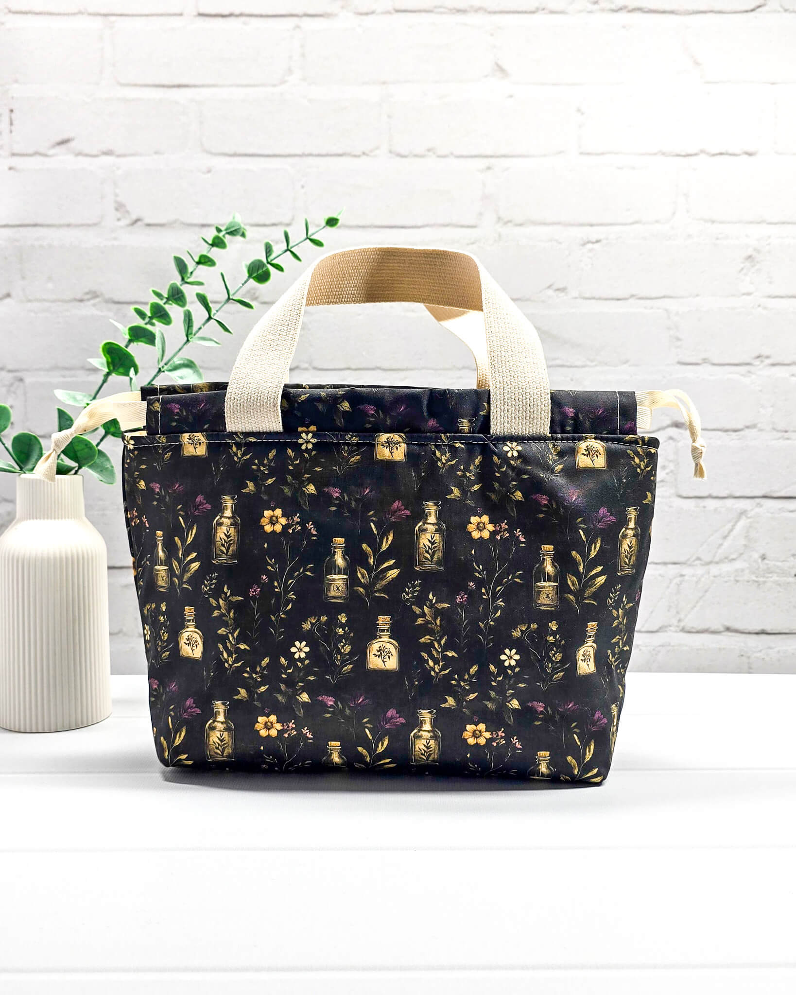 Projekttasche Alva S Secret Garden mit zwei hellen Griffen und dunklem botanischem Muster mit Blüten und kleinen goldenen Fläschchen. Handgenäht in Bayern von NadelMikado.