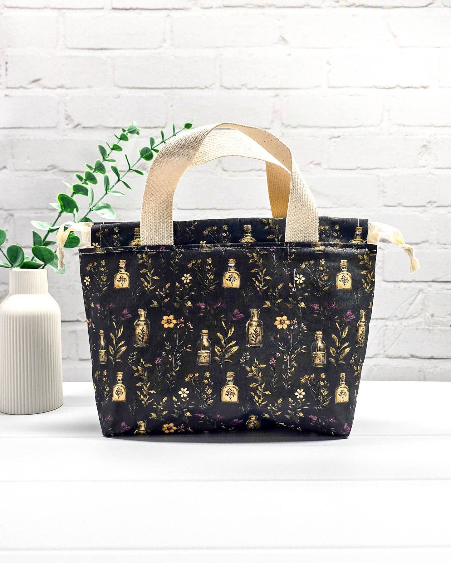 Projekttasche Alva S Secret Garden mit zwei hellen Griffen und dunklem botanischem Muster mit Blüten und kleinen goldenen Fläschchen. Handgenäht in Bayern von NadelMikado.