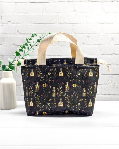 Projekttasche Alva S Secret Garden mit zwei hellen Griffen und dunklem botanischem Muster mit Blüten und kleinen goldenen Fläschchen. Handgenäht in Bayern von NadelMikado.