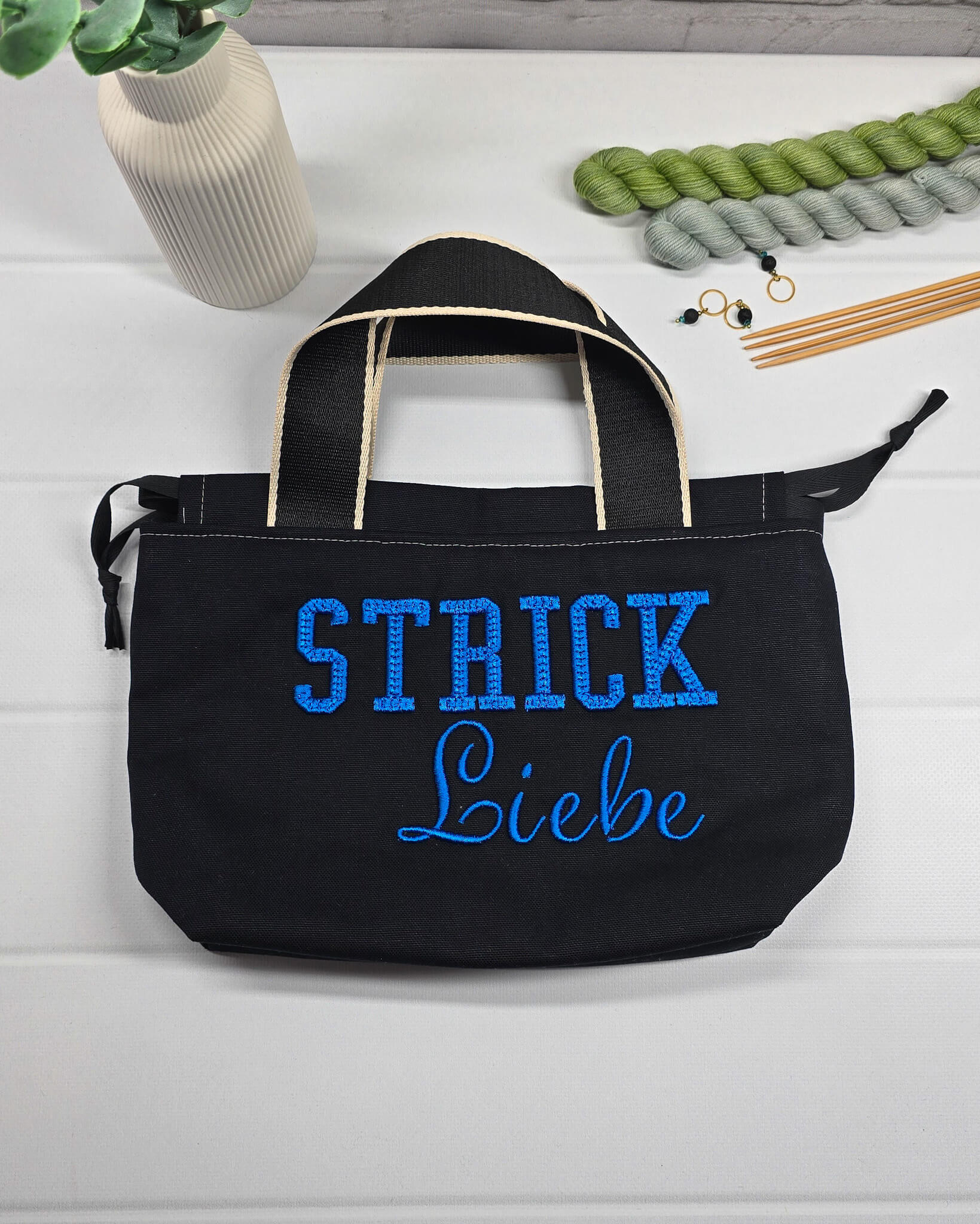 Projekttasche Alva S aus schwarzem Canvas mit blauer 3D-Stickerei Strickliebe, zwei Henkeln und schwarzem Kordelzugkragen. Handgenäht in Bayern von Nadelmikado.