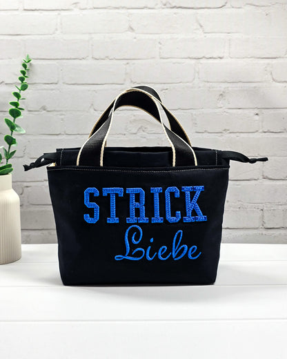 Projekttasche Alva S aus schwarzem Canvas mit blauer 3D-Stickerei Strickliebe, zwei Henkeln und schwarzem Kordelzugkragen. Handgenäht in Bayern von Nadelmikado.