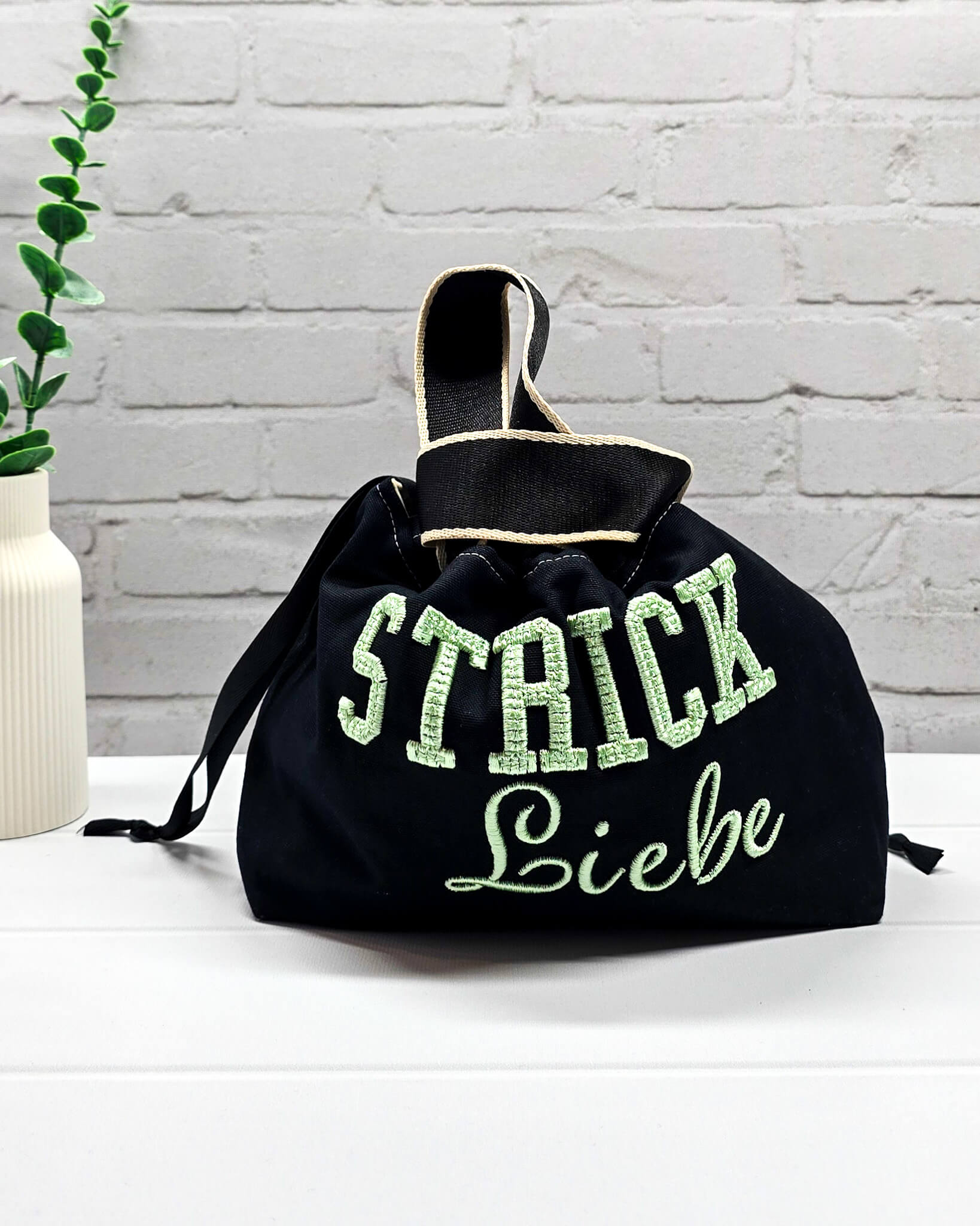 Projekttasche Alva S aus schwarzem Canvas mit mintgrüner 3D-Stickerei Strickliebe, Henkeln und schwarzem Kordelzugkragen. Handgenäht in Bayern von Nadelmikado.