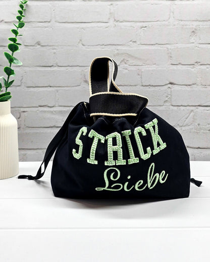 Projekttasche Alva S aus schwarzem Canvas mit mintgrüner 3D-Stickerei Strickliebe, Henkeln und schwarzem Kordelzugkragen. Handgenäht in Bayern von Nadelmikado.