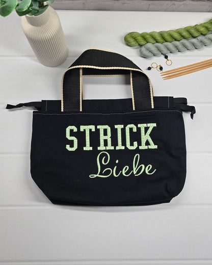 Projekttasche Alva S aus schwarzem Canvas mit mintgrüner 3D-Stickerei Strickliebe, Henkeln und schwarzem Kordelzugkragen. Handgenäht in Bayern von Nadelmikado.