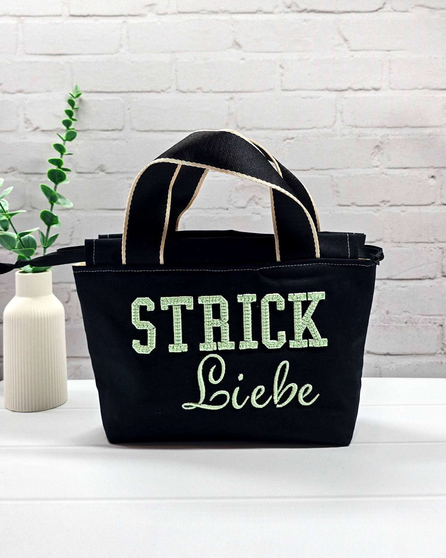Projekttasche Alva S aus schwarzem Canvas mit mintgrüner 3D-Stickerei Strickliebe, Henkeln und schwarzem Kordelzugkragen. Handgenäht in Bayern von Nadelmikado.