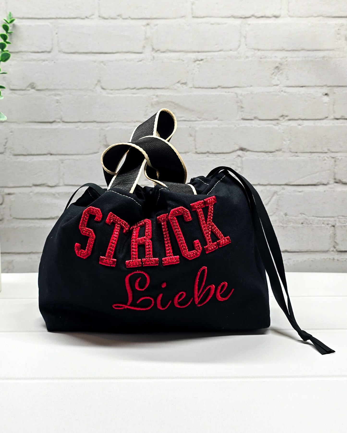 Projekttasche Alva S aus schwarzem Canvas mit rot gesticktem 3D-Schriftzug Strickliebe, Henkeln und Kordelzugkragen. Handgenäht in Bayern von Nadelmikado.