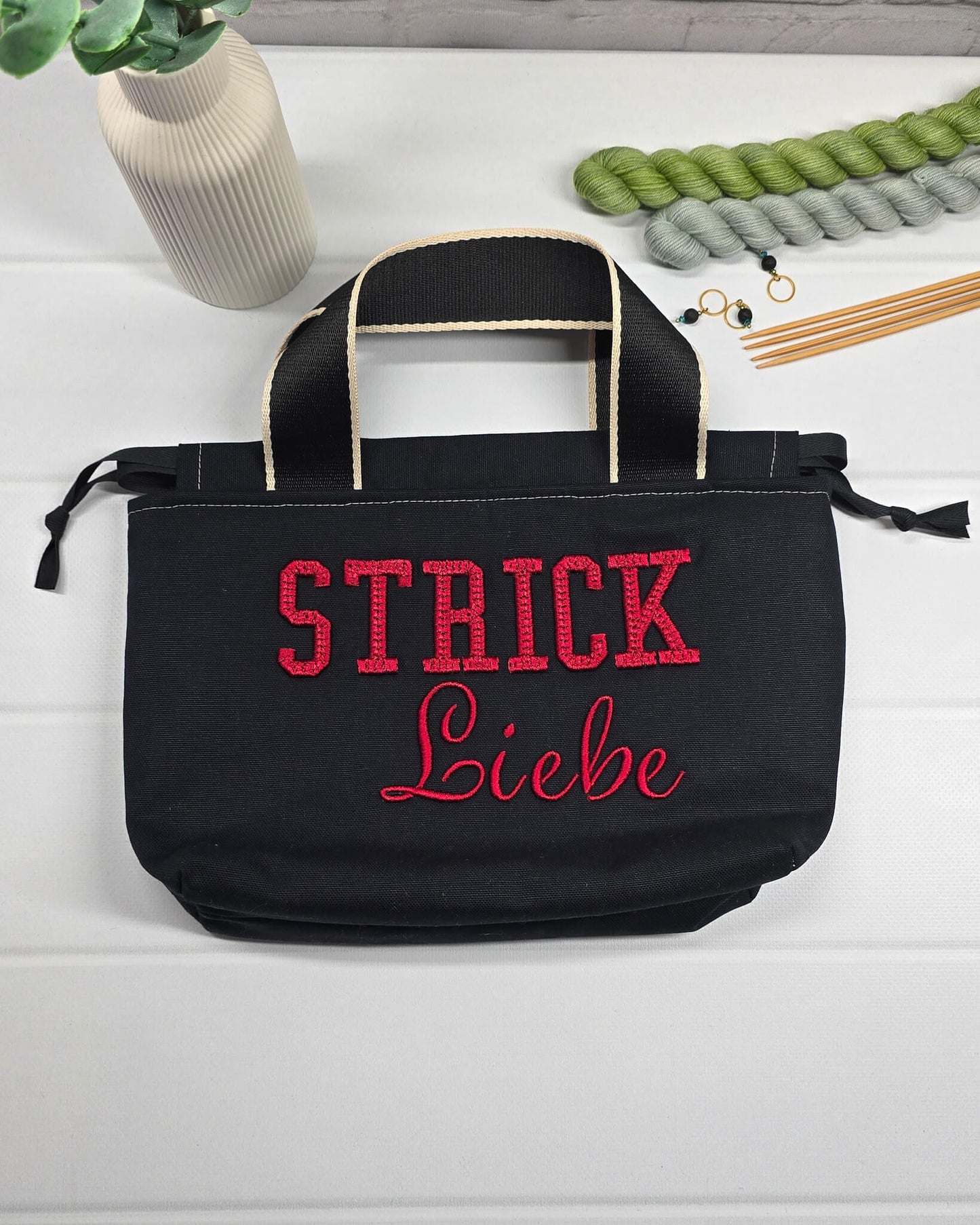 Projekttasche Alva S aus schwarzem Canvas mit rot gesticktem 3D-Schriftzug Strickliebe, Henkeln und Kordelzugkragen. Handgenäht in Bayern von Nadelmikado.