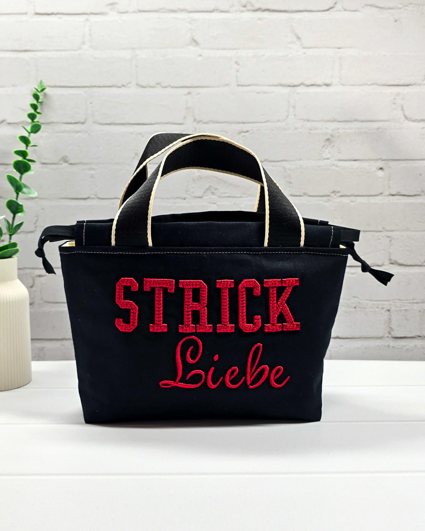 Projekttasche Alva S aus schwarzem Canvas mit rot gesticktem 3D-Schriftzug Strickliebe, Henkeln und Kordelzugkragen. Handgenäht in Bayern von Nadelmikado.