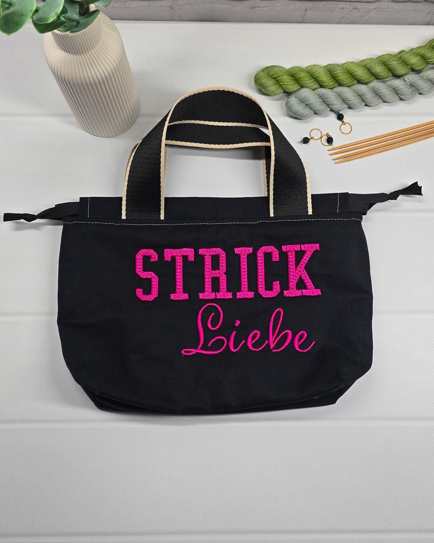 Projekttasche Alva S aus schwarzem Canvas mit erhabenem 3D-Stickzug Strickliebe in Pink, Henkeln und Kordelzugkragen. Handgenäht in Bayern von Nadelmikado.